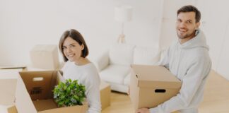 Positive female and male property owners pose with personal belongings in carton boxes.